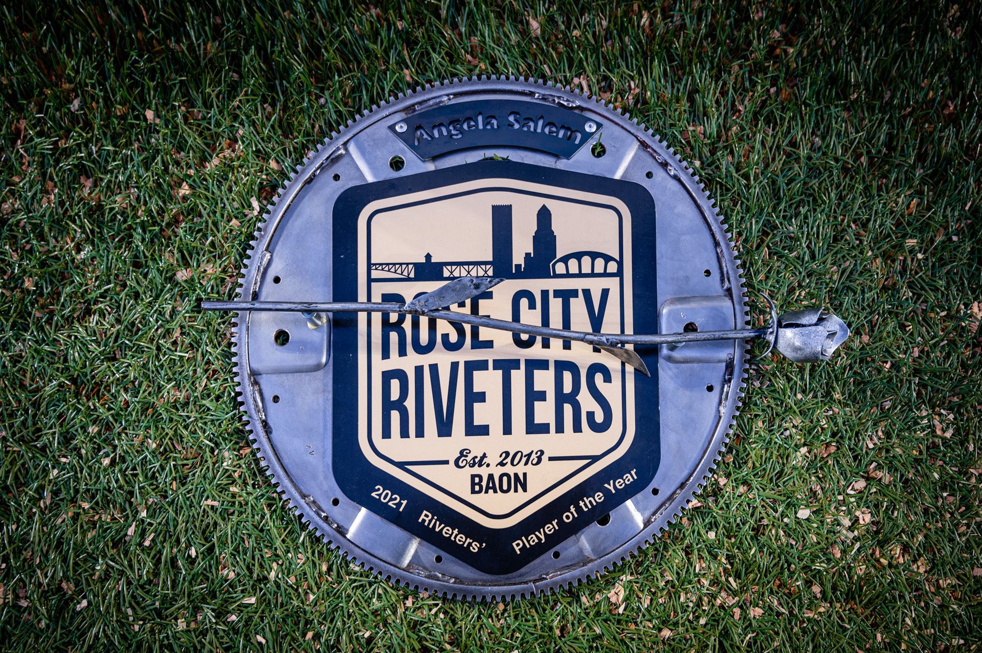 Trophy is a large circular gear with a plaque in the center with the Rose City Riveters crest reading 2021 Riveters' Player of the Year. Angela Salem's name is at the top. There is a metal rose attached that runs across the crest. 