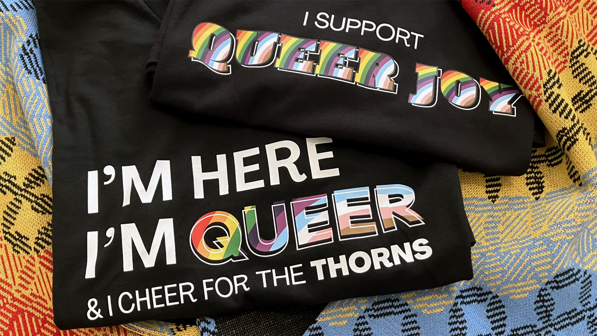 Two black t-shirts. One design reads "I support queer joy" and the other reads "I'm here, I'm queer, & I cheer for the Thorns". The text is in pride colors for the words queer and joy.